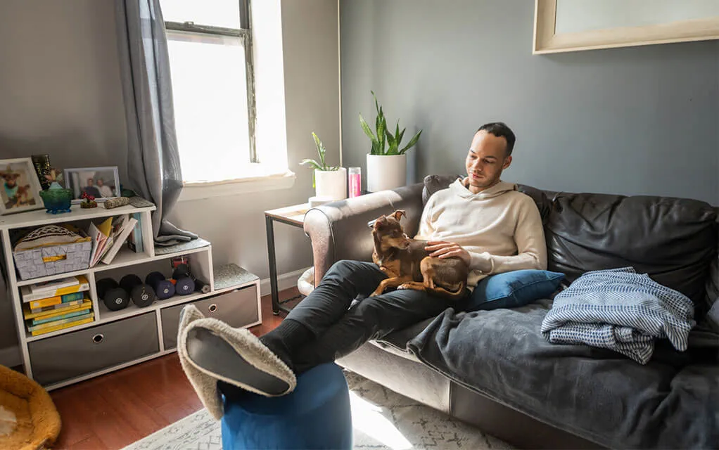 A man sits on his couch with his feet up on an ottoman with his small dog in his lap, smiling, as sunlight streams into the room from the window
