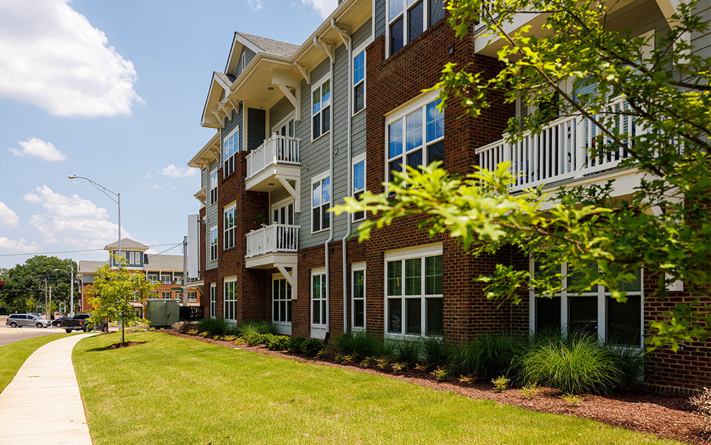 Outside view of a brick and vinyl facade of an apartment complex from the sidewalk of a busy street as some clouds are in the sky on a bright and sunny summer day