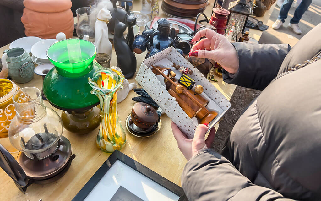 A woman in a winter jacket stands at a table with items all over it for sale, holding a box with a wooden figurine inside of it