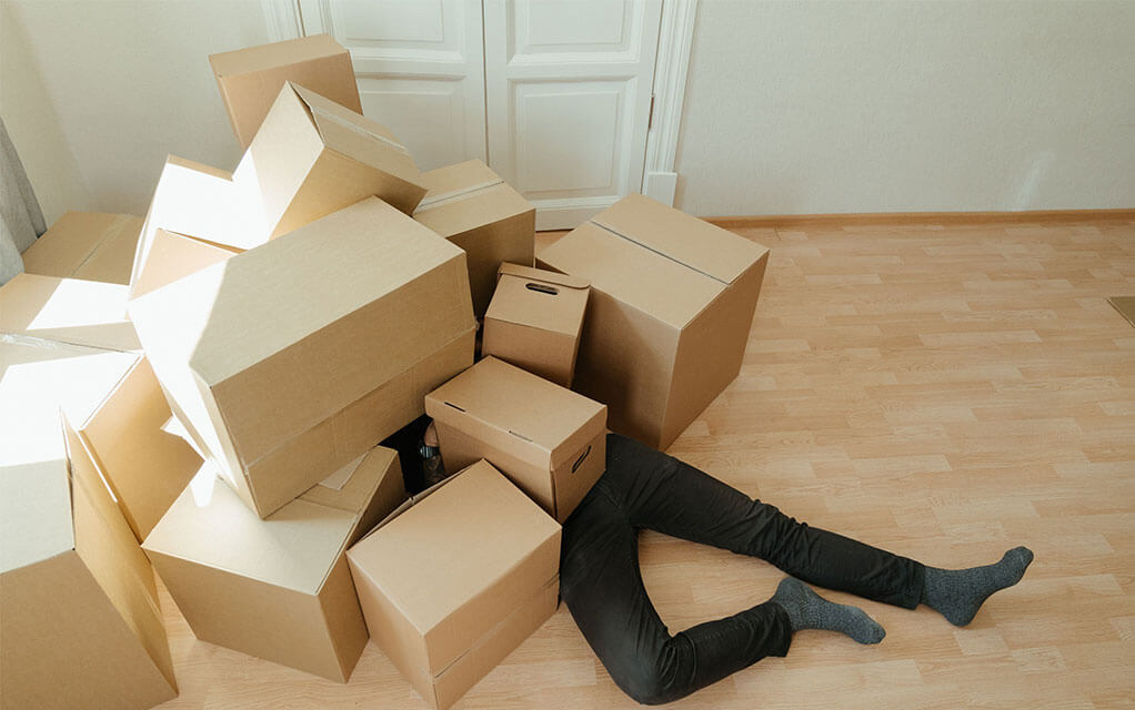 A person sits under a pile of cardboard boxes covering their torso on up while their legs are splayed out on the floor as sunlight streams into the room