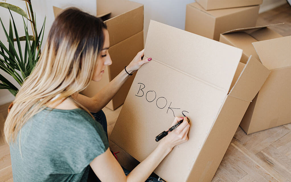 A young woman writes the word "BOOKS" on an empty cardboard box, as other moving boxes that are open and waiting to be packed and secured sit in the background