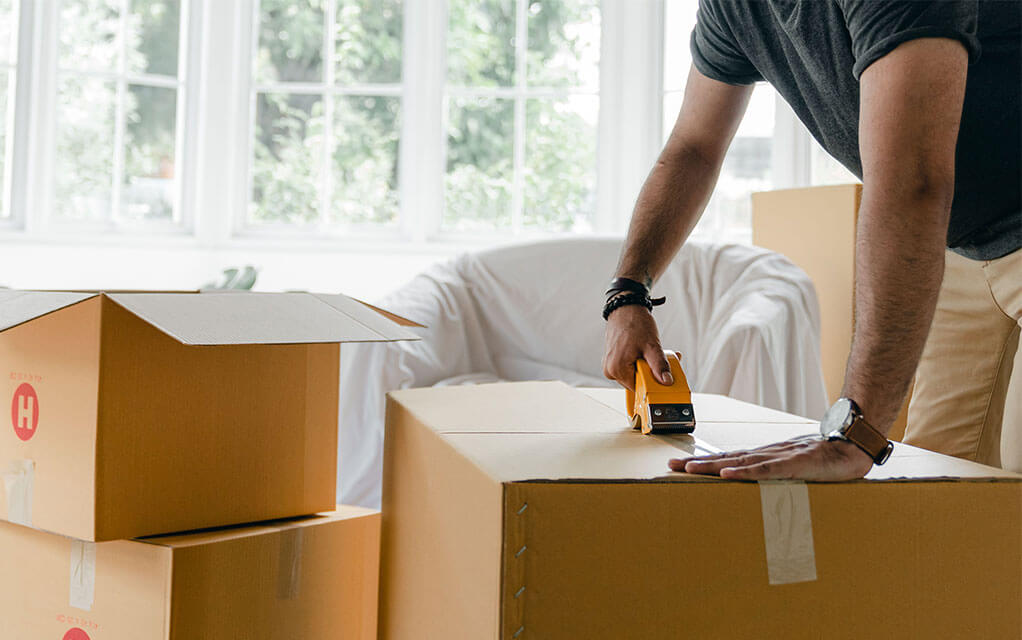 A man is seen taping up the top of a large box with a tape gun as other open boxes sit on the floor waiting to be taped