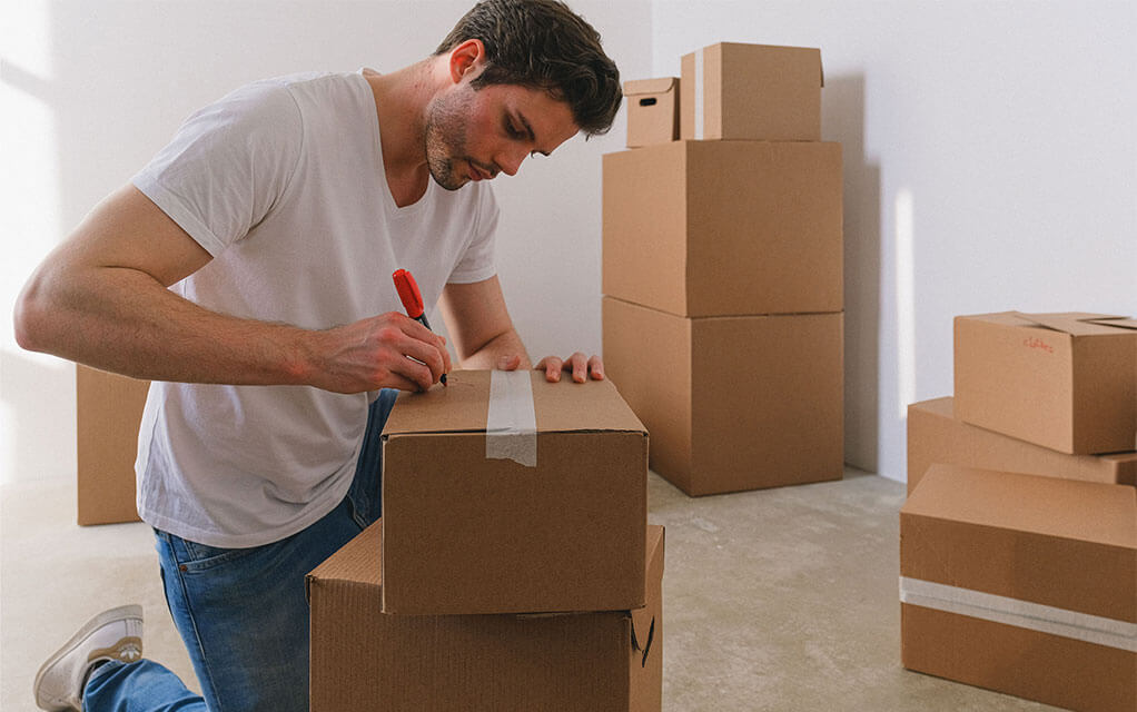 A man kneels down in front of a couple of stacked boxes as he begins to write something on the top of the taped up box with a red magic marker, as more taped and packed boxes sit around the room
