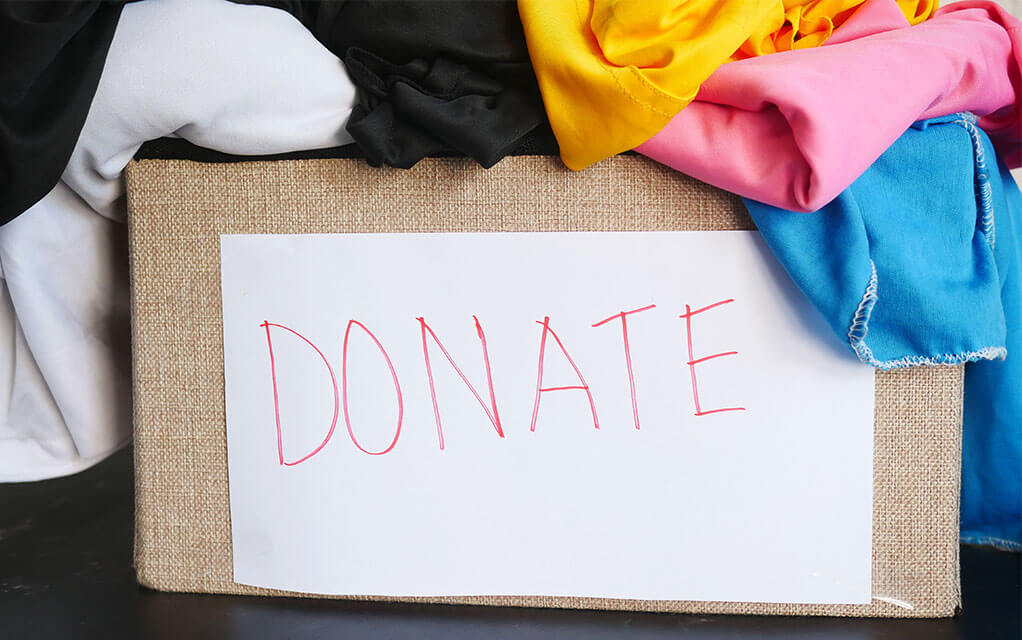 A box with the word "DONATE" written on it with red ink sits on a table with clothing overflowing out of it