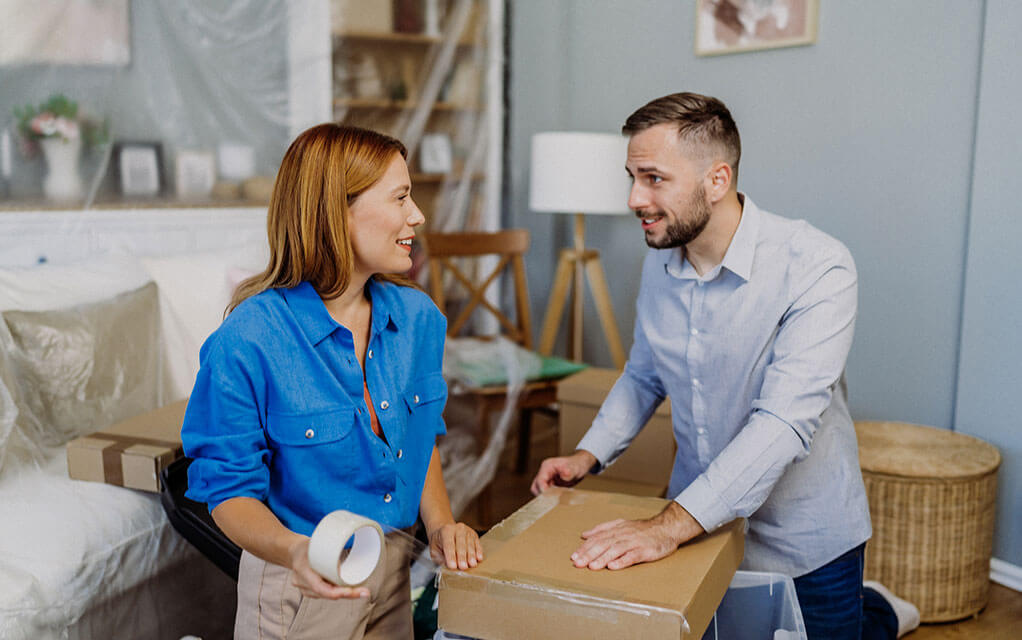 A young couple packs boxes in their home as their belongings lay around them in their home