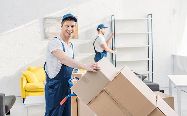 Two professional movers are packing up an apartment with the laborer in the foreground smiling as he moves a hand truck loaded with boxes