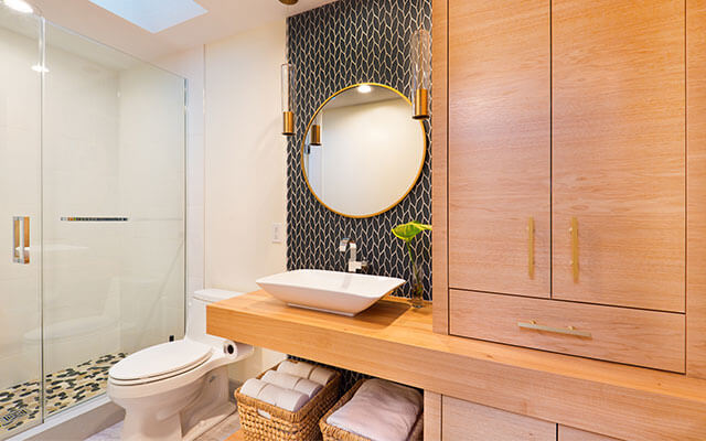 Up close view of a upscale home bathroom sink with a circular mirror, with a large storage cabinet next to the sink, with towels in baskets on a hanging shelf underneath