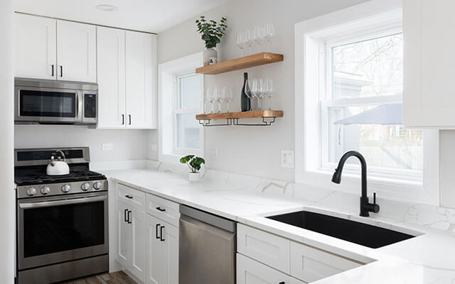 The view of a home's kitchen that is almost entirely white in color, with floating shelves present on the wall holding a plant, wine glasses, and a bottle of wine.
