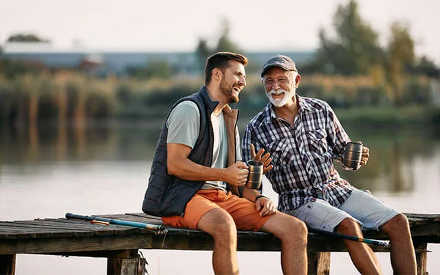 An older man and his son sit on the dock of a lake with fishing rods sitting next to them, laughing and enjoying the nice day outside