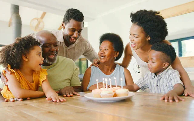 A woman sits surrounded by her family with cupcakes adorned with lit candles as several generations of family celebrate the woman's birthday