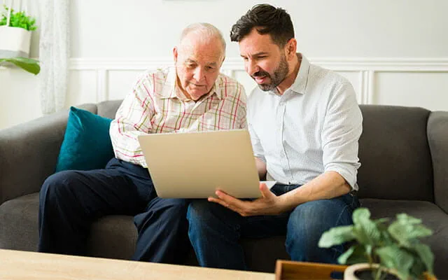 An elderly man and his son sit on a couch in a home as the son walks through something with his father on his laptop
