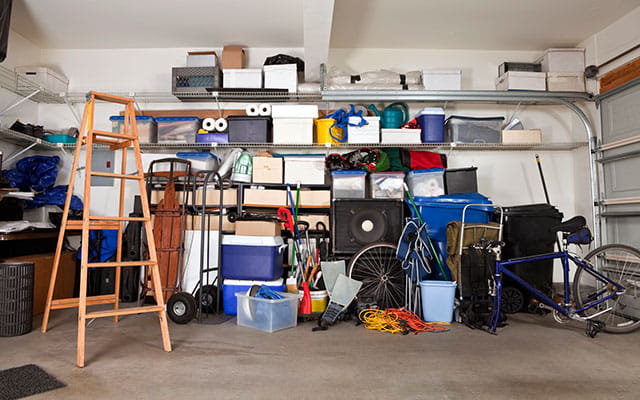 A bunch of random bins, boxes, and stuff sits along the inside wall of a closed garage with a basic ladder open next the the items