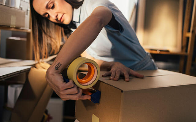 A woman uses a tape roller to begin taping shut a brown cardboard box filled with items inside of her garage